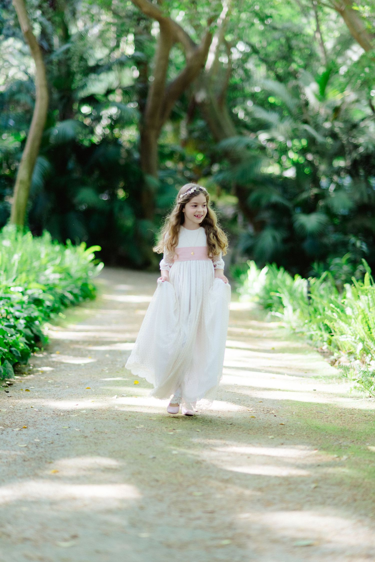 Young girl in white and pink communion dress walking along a lush garden path.