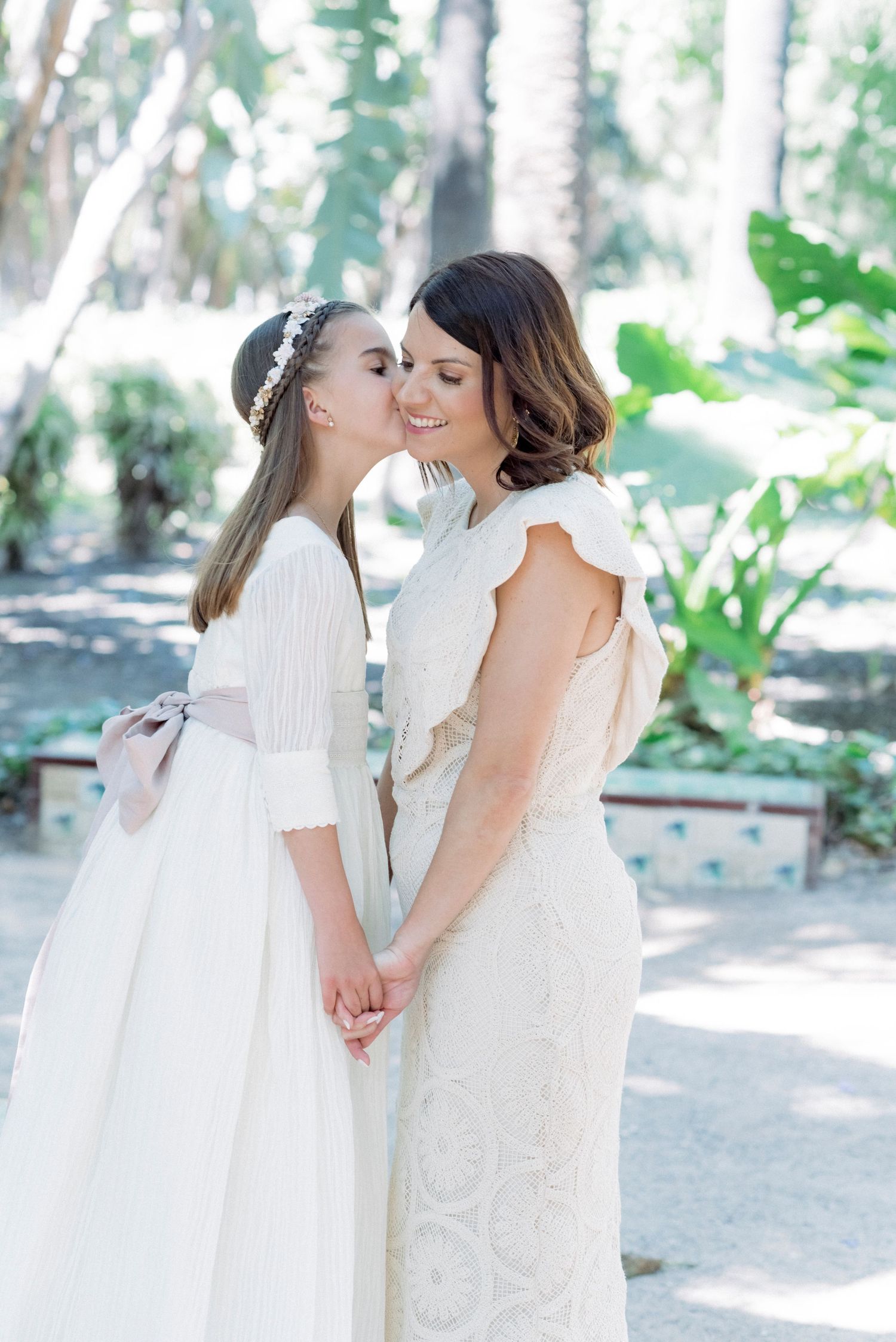 A bride and young flower girl in white dresses share a tender moment holding hands in a sunlit garden.