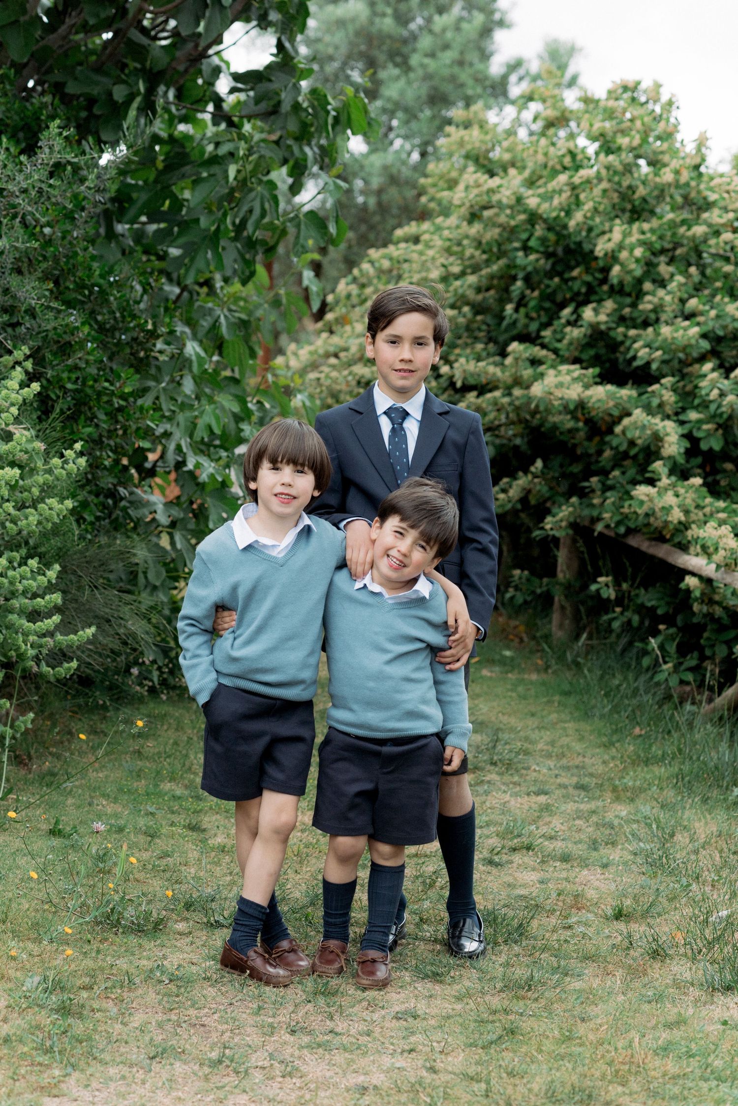 Three boys in formal attire posing together in a lush green garden setting.