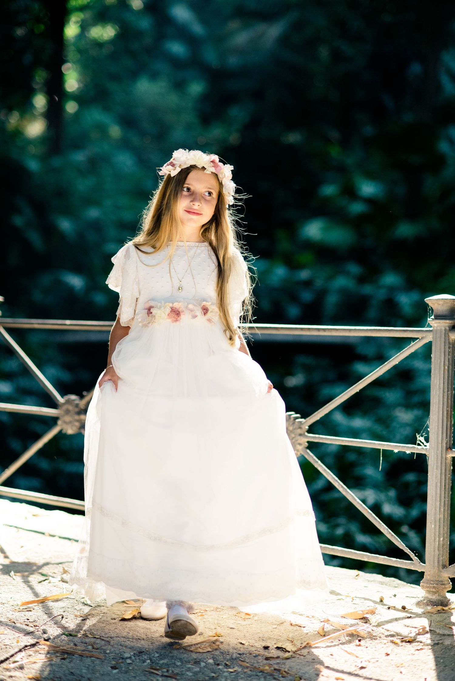 Young girl in white dress and flower crown stands on sunlit garden bridge.