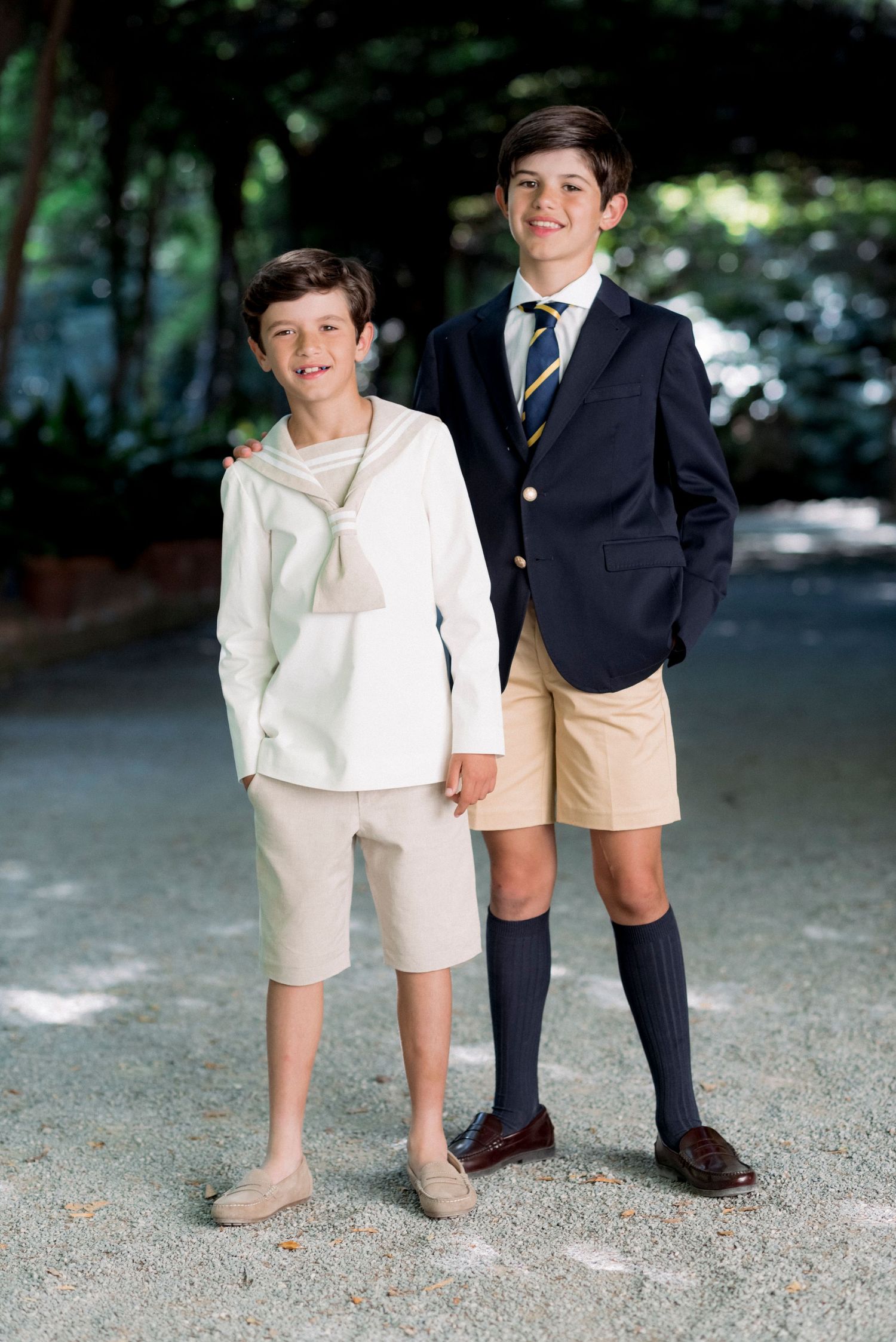 Two smiling boys in formal communion outfits posing together on a garden path.