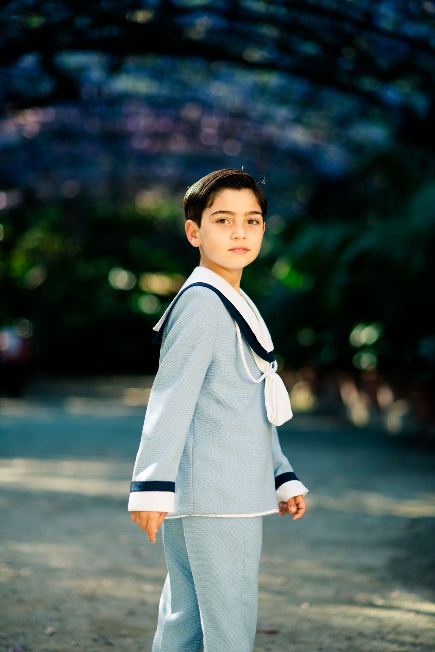 Boy in light blue sailor suit standing in garden path.