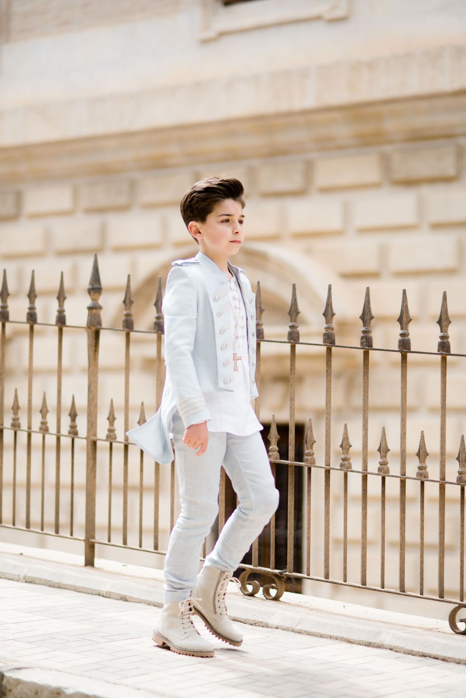 A boy in a white communion suit and boots stands confidently by an iron fence.