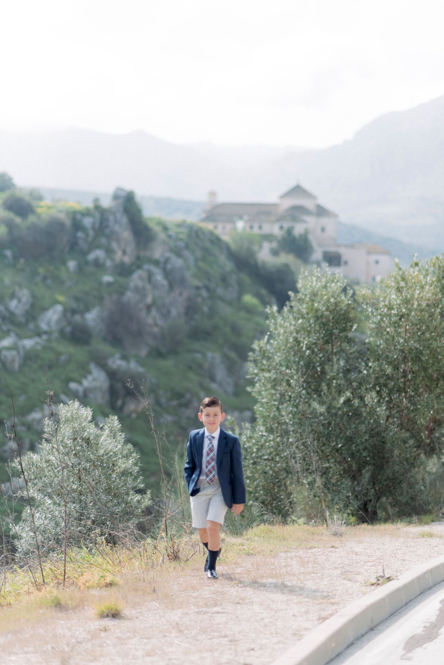 A well-dressed boy in a navy blazer walks along a scenic path with mountains and a church in the background.