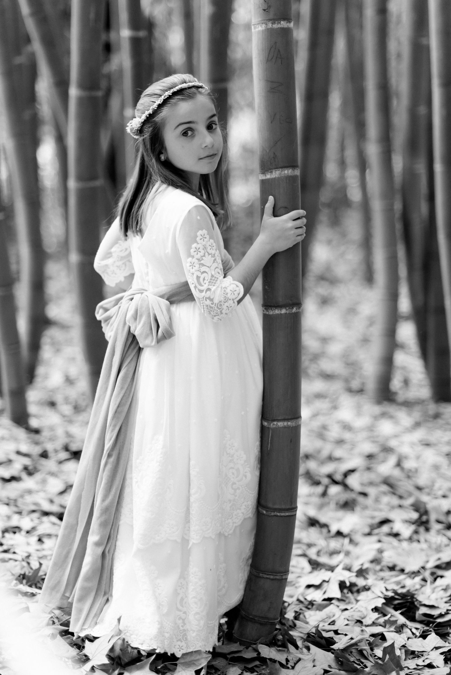 Girl in white lace communion dress with bow holds bamboo tree in black and white forest photo.