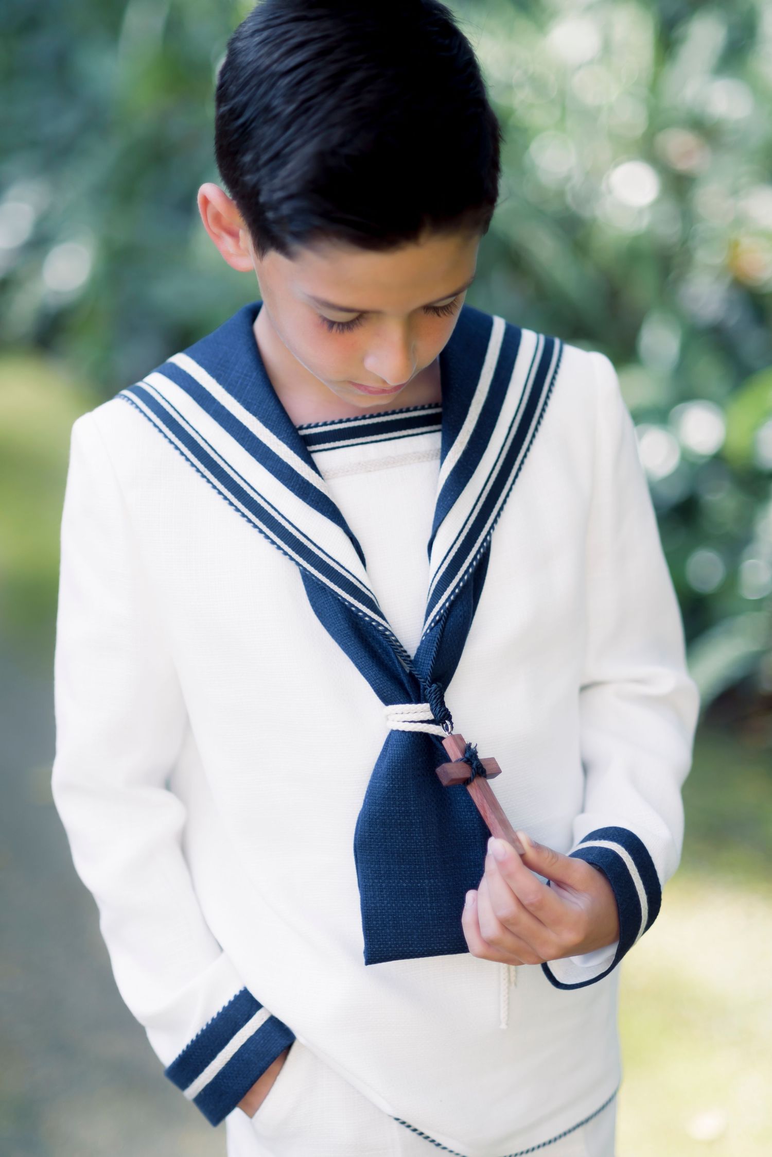 Boy in white and navy sailor communion suit holds rosary in lush garden setting.