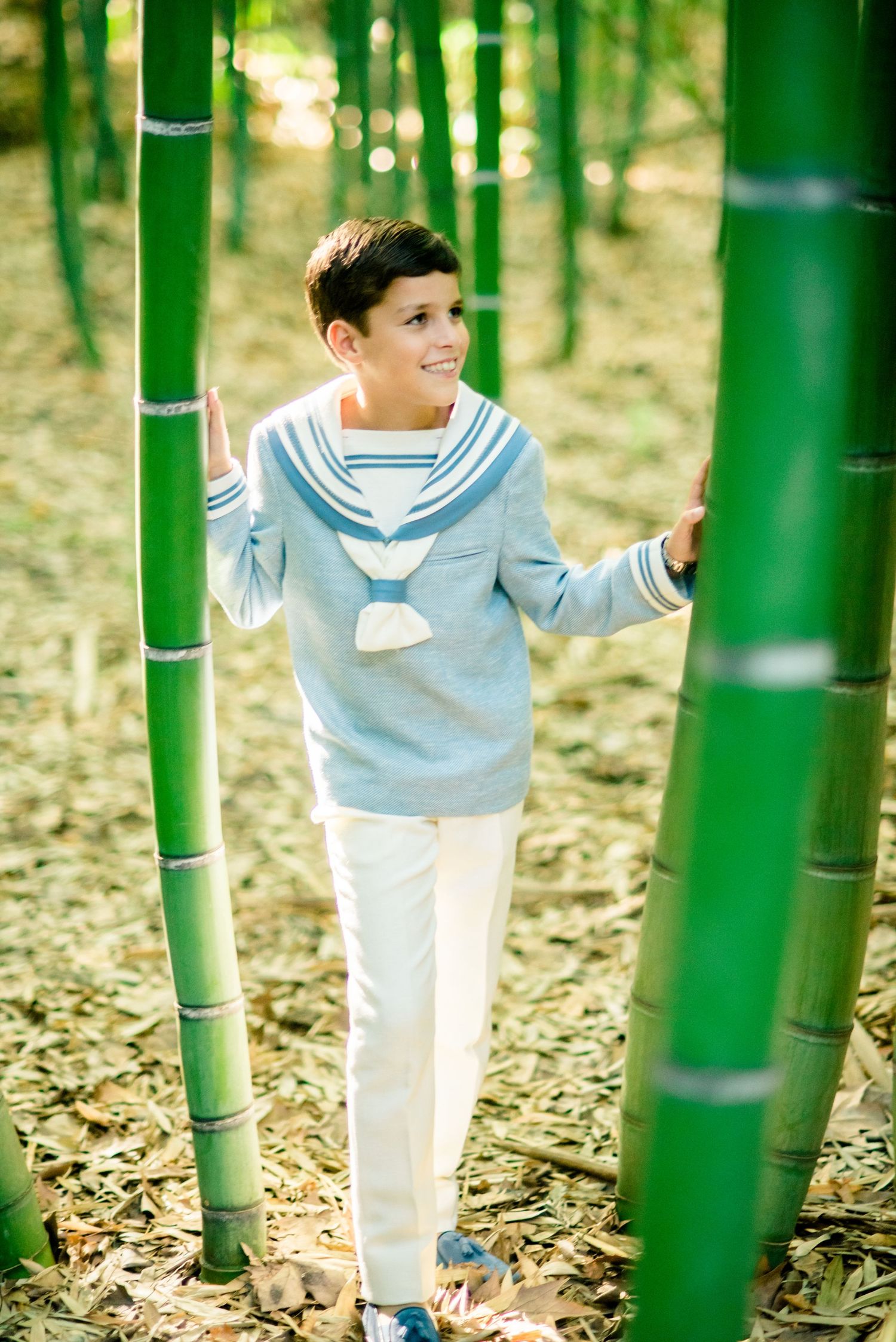 Smiling boy in sailor-style blue top walking through tall green bamboo forest.
