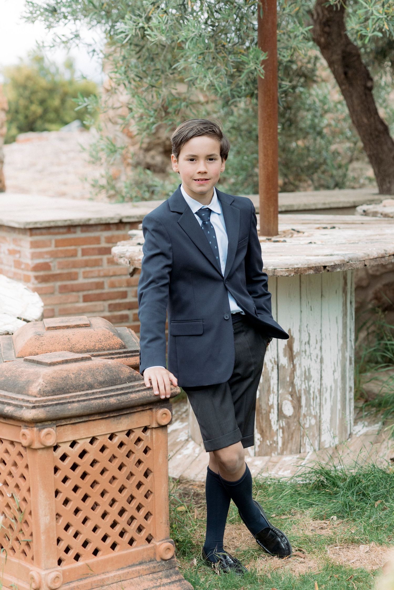 Boy in navy suit jacket, tie, and shorts with knee-high socks poses outdoors near brick structure.