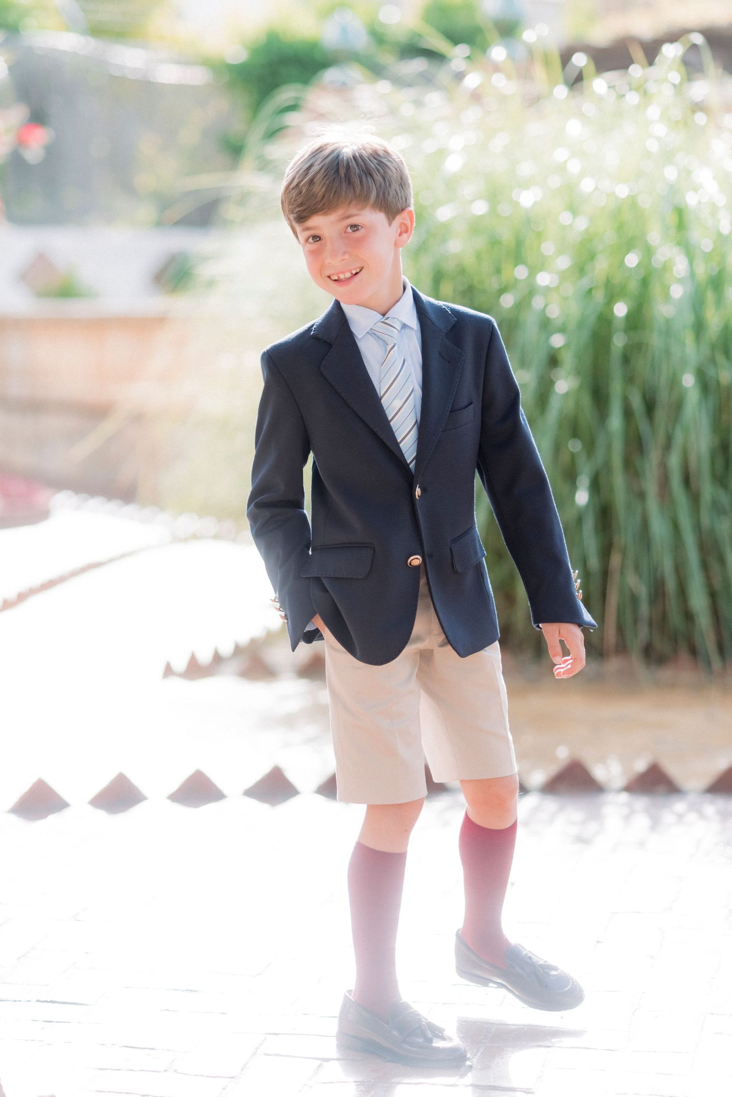 Smiling boy in navy blazer, striped tie, and khaki shorts posing outdoors.