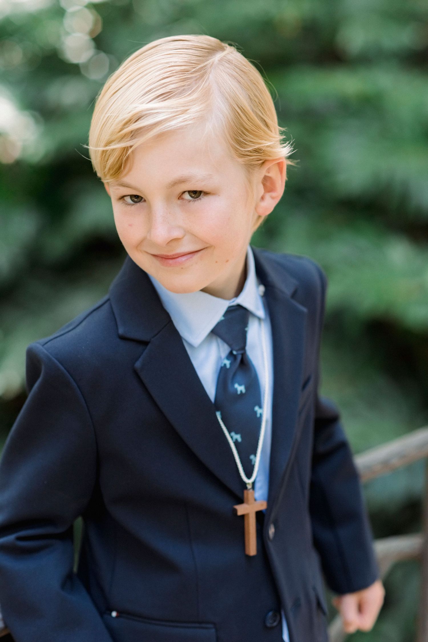 Blonde boy in navy suit with cross necklace and tie smiles outdoors among green foliage.