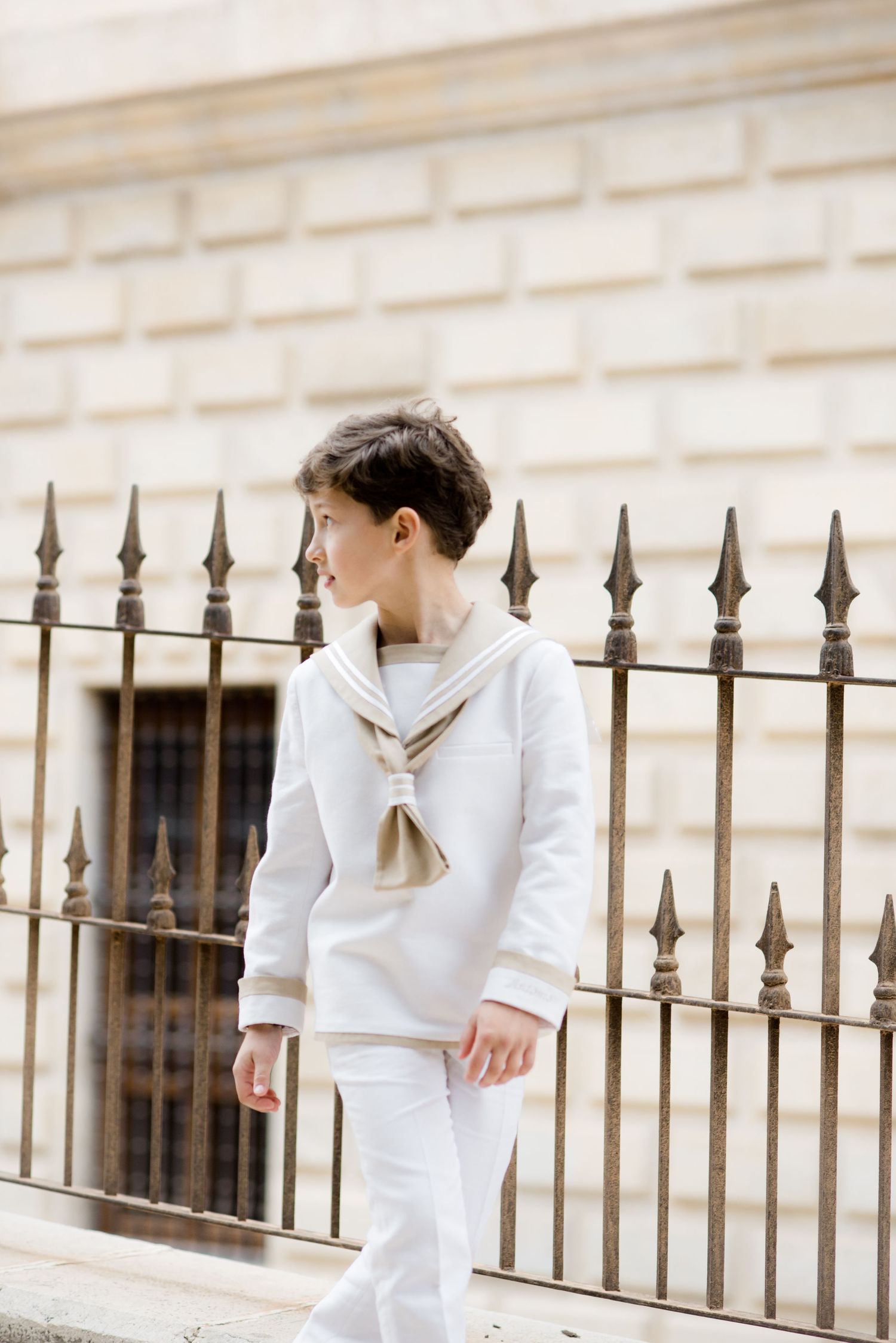 Boy in white sailor suit standing by iron fence.