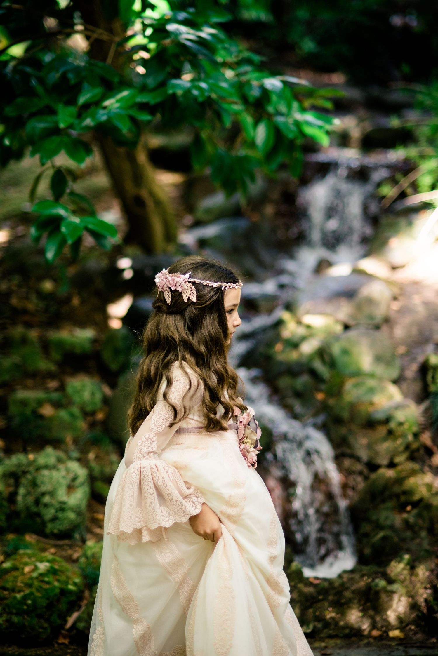 Girl in white lace communion dress with floral crown standing near garden waterfall.
