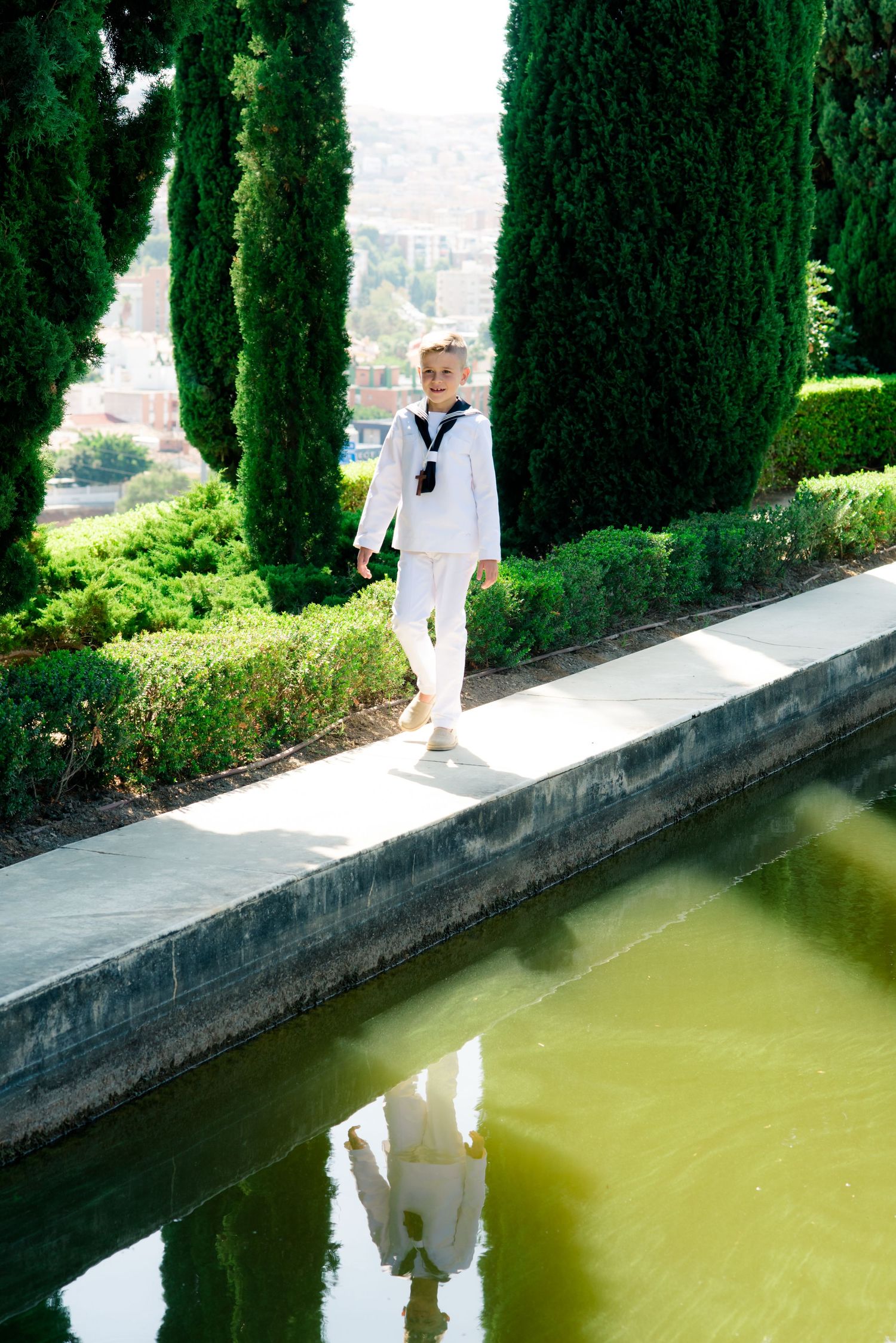 Child in white sailor outfit walks along garden pool edge lined with tall cypress trees.