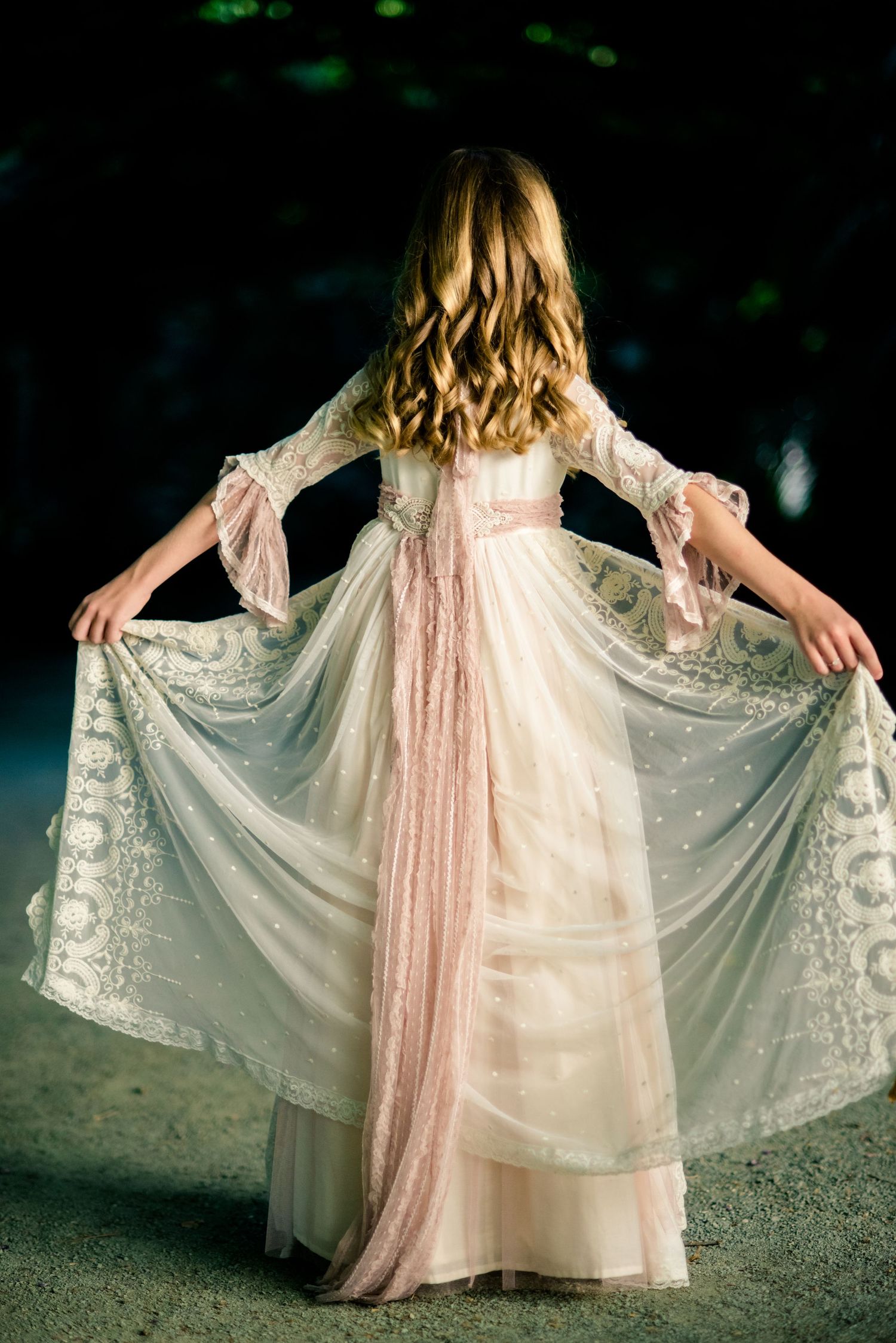 Back view of girl in flowing cream and pink lace dress spreading skirt in dark garden.