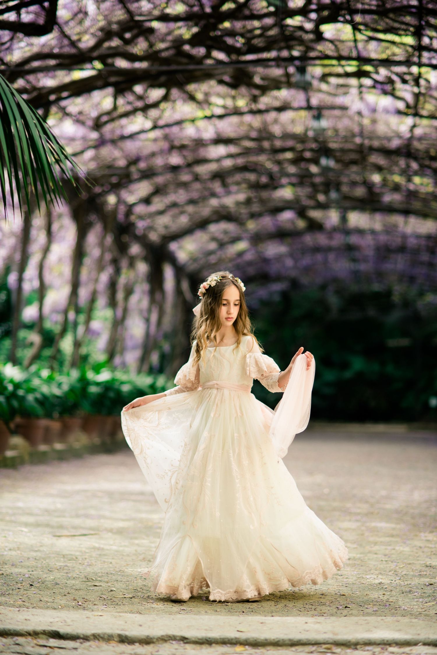 Girl in flowing cream dress twirling under wisteria archway.
