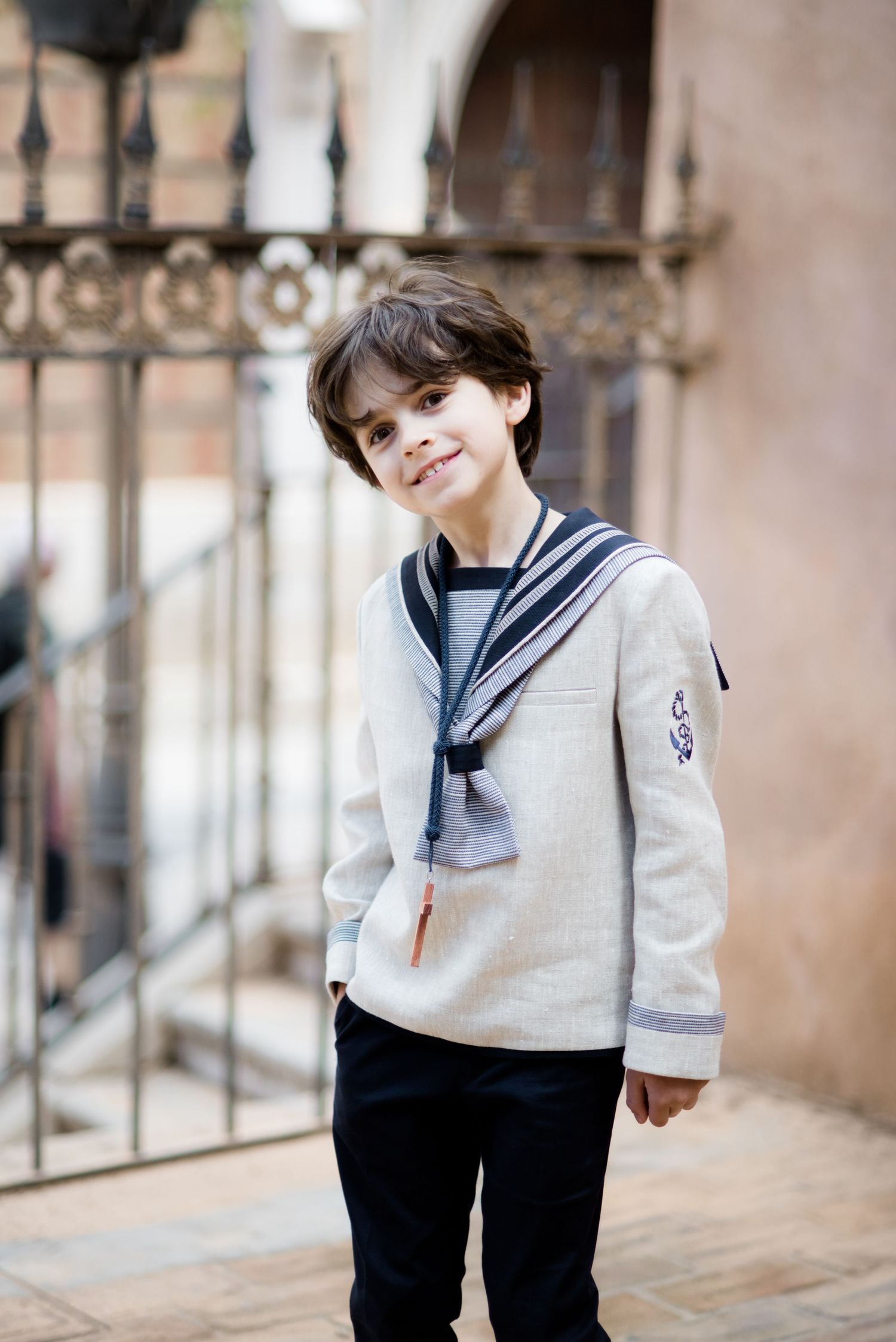 A smiling boy in a sailor-style communion outfit stands near an ornate iron gate.