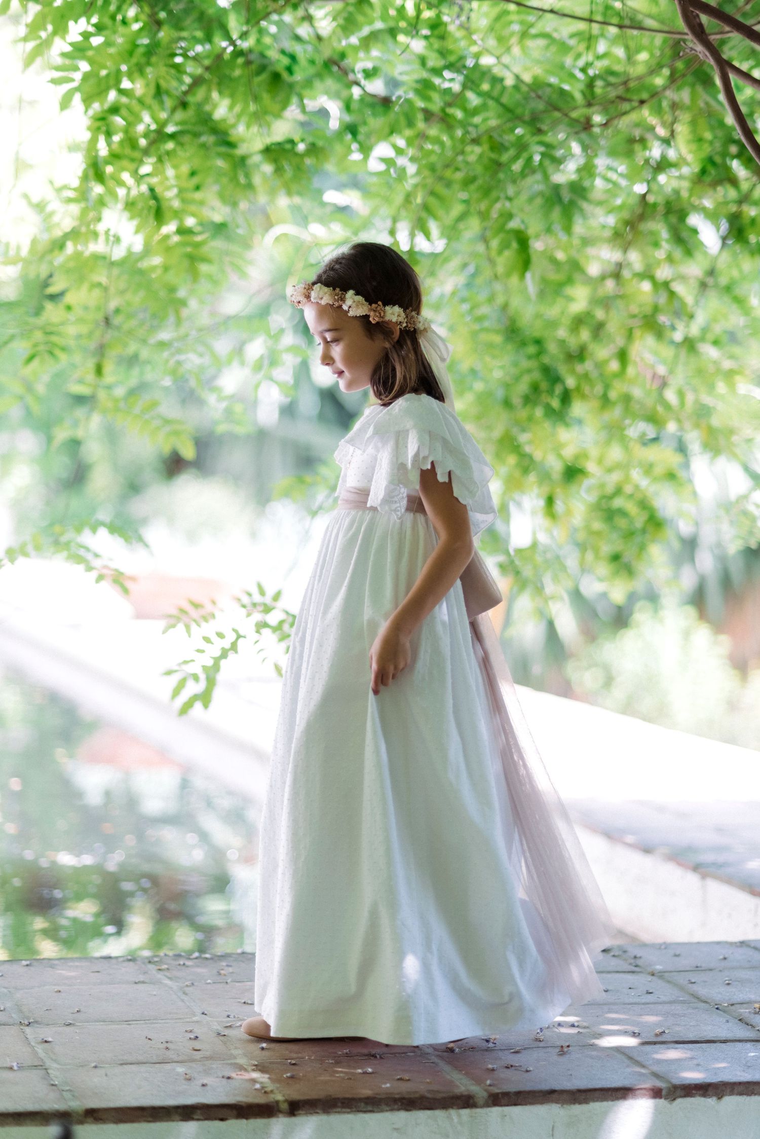 Girl in elegant white communion gown with floral crown standing under green tree canopy.