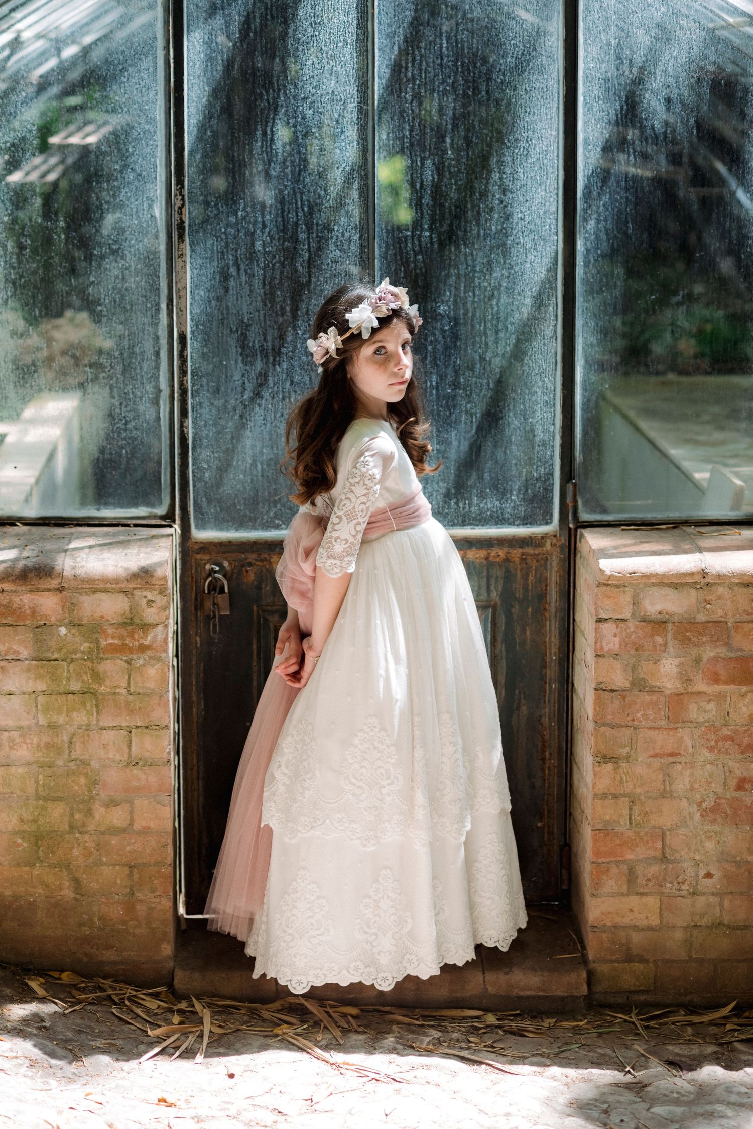 Girl in lace communion dress with pink sash and floral crown posing by vintage glass door.