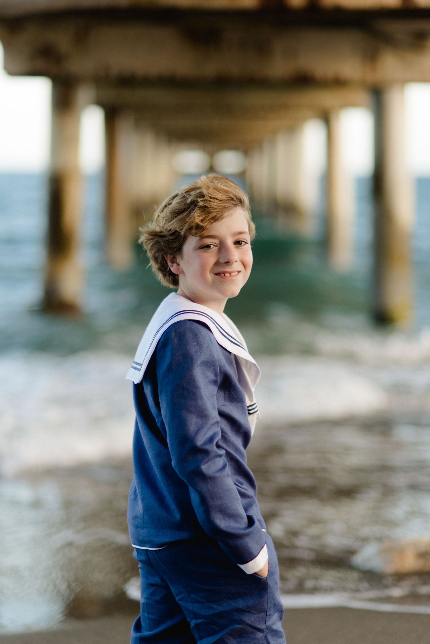 Smiling boy in navy sailor suit standing on beach under a wooden pier.