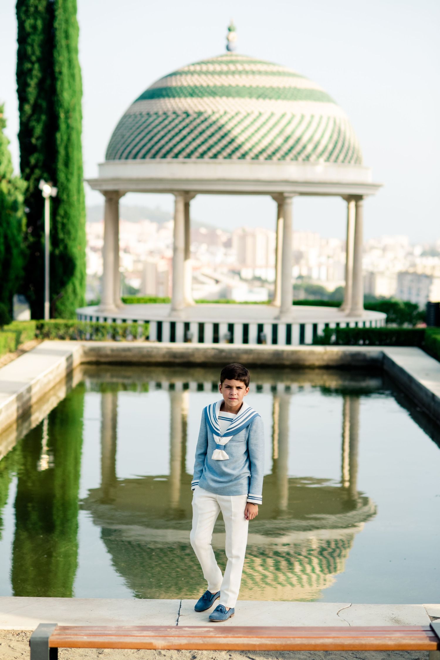 Boy in blue sailor outfit standing by ornate domed pavilion reflected in garden pool.