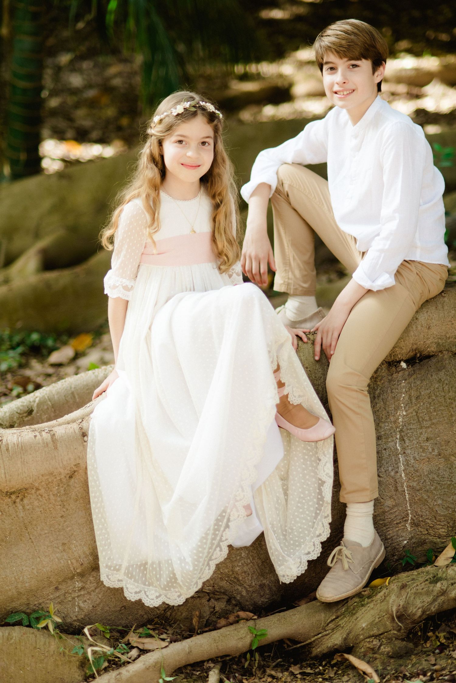 Two children in formal white communion attire sit together on a stone ledge outdoors.