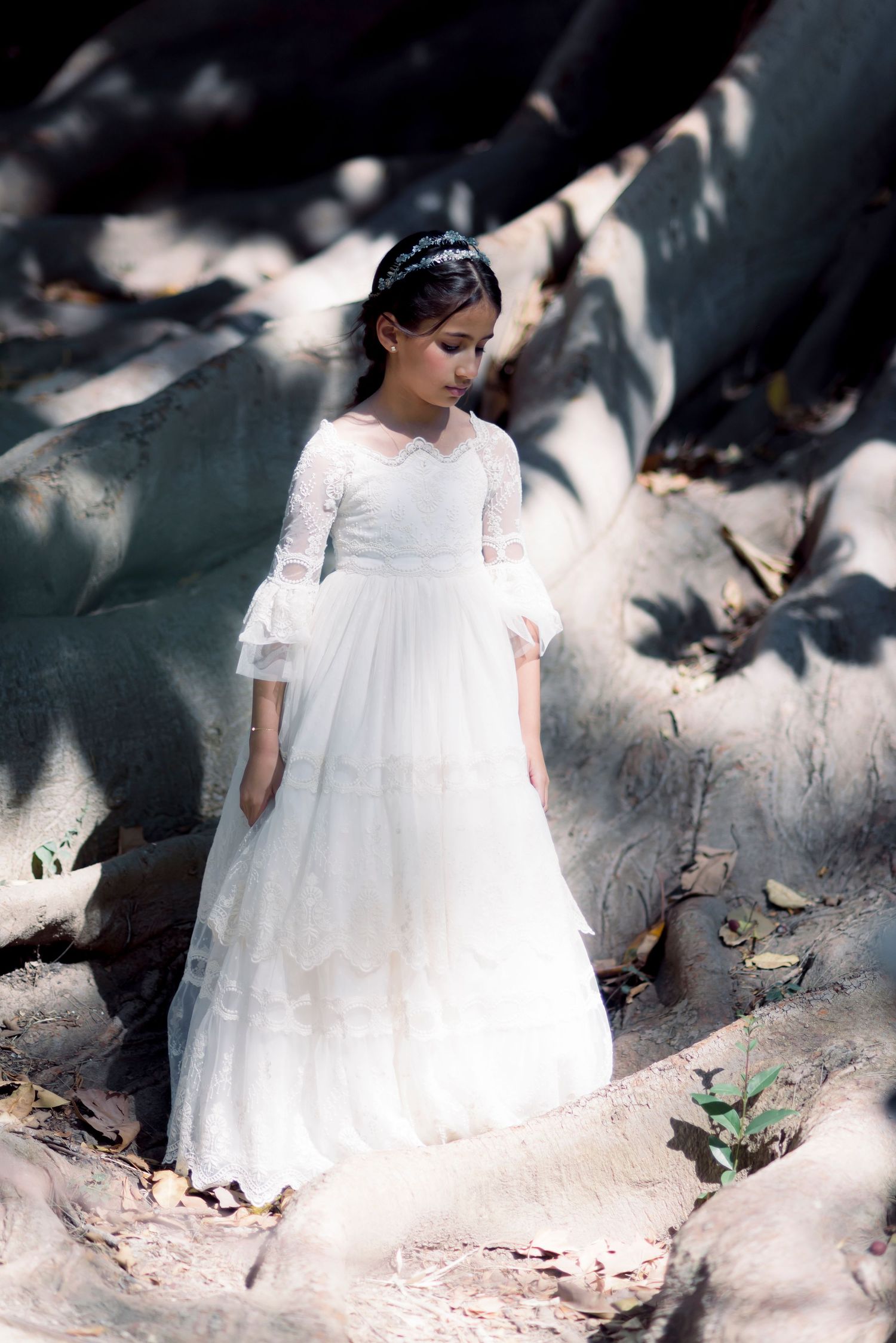 Girl in white lace First Communion dress stands among large tree roots in sunlight.