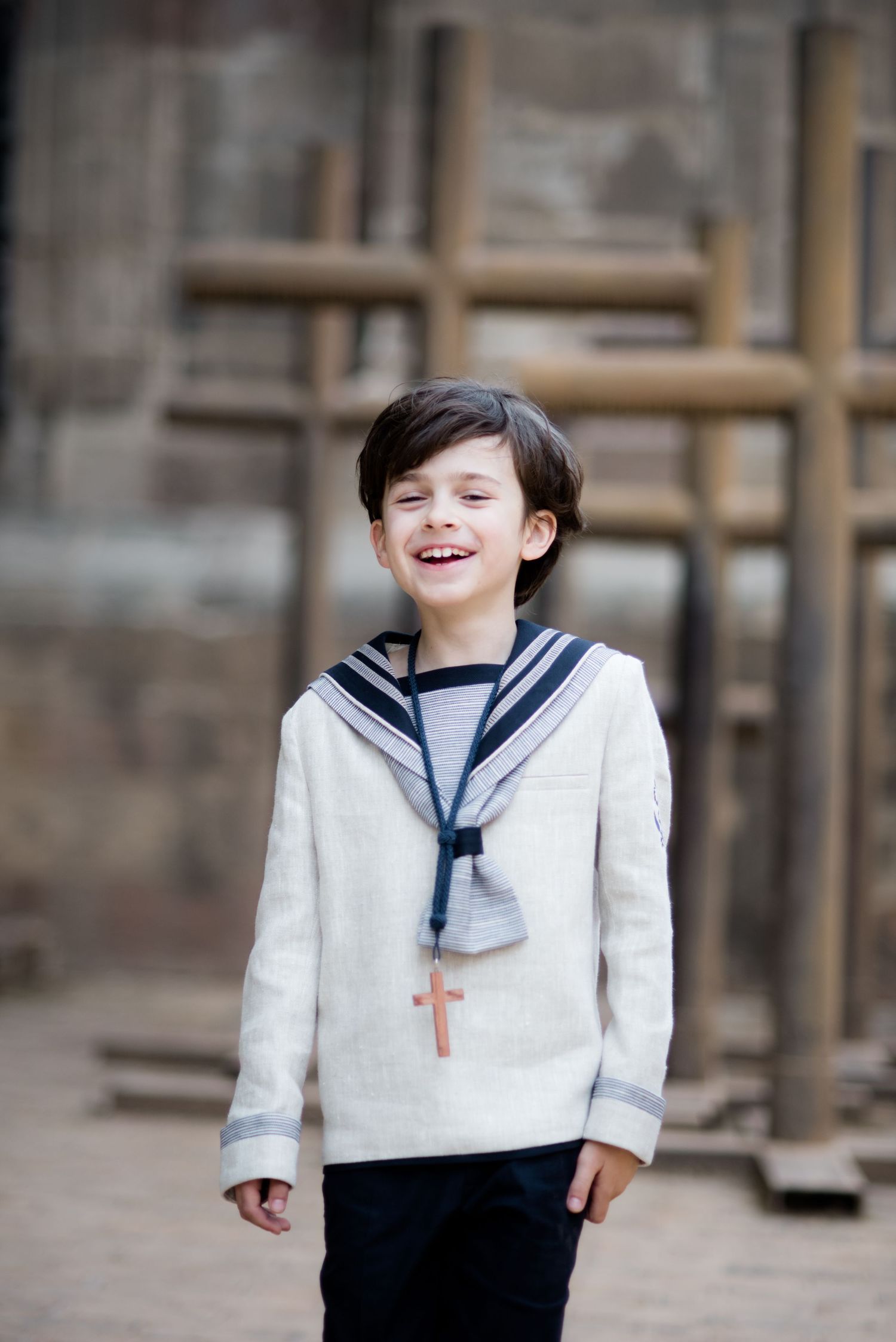A laughing boy in a sailor communion suit with a cross necklace poses outdoors.