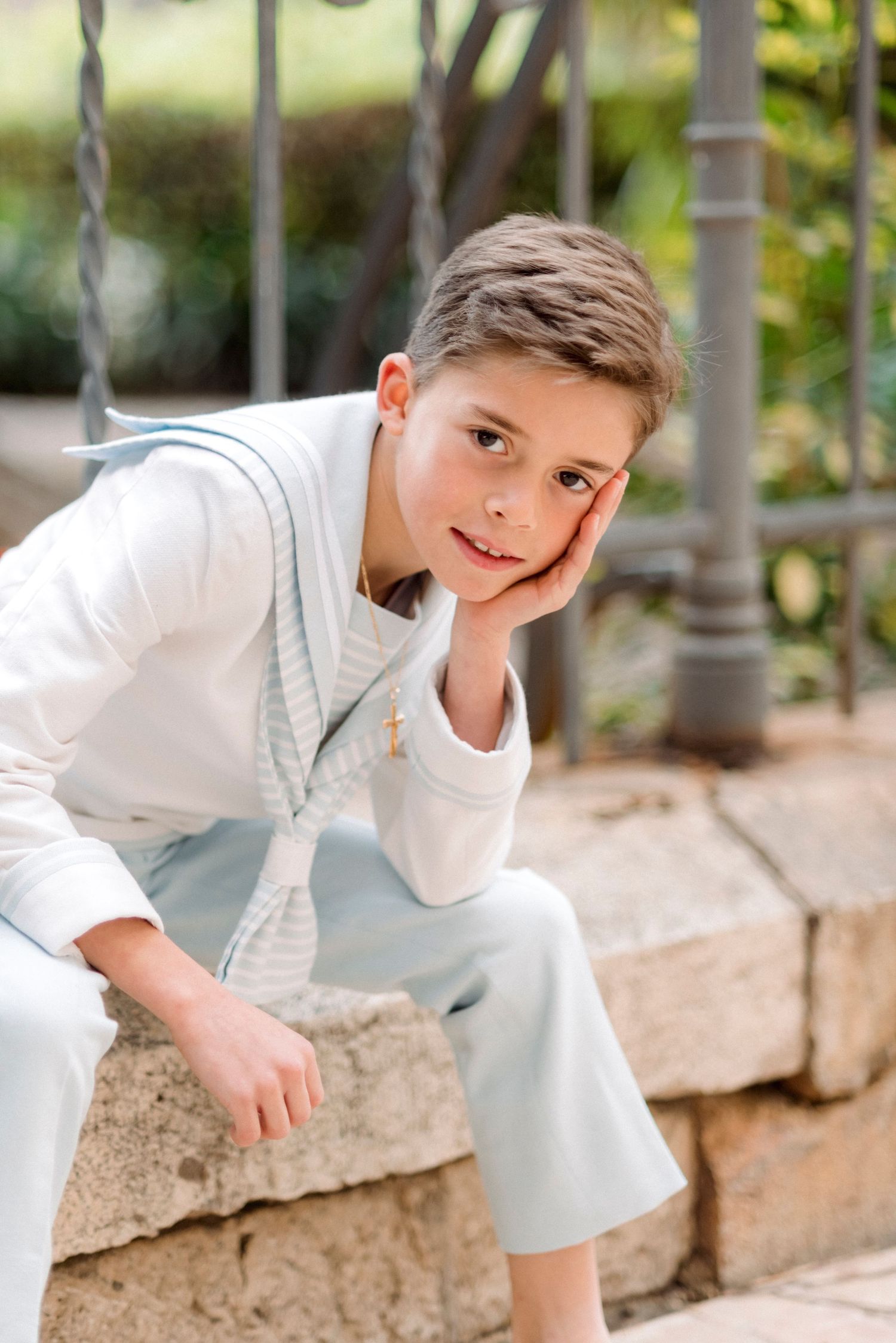 Boy in white communion outfit with blue vest sits on stone ledge resting cheek on hand.