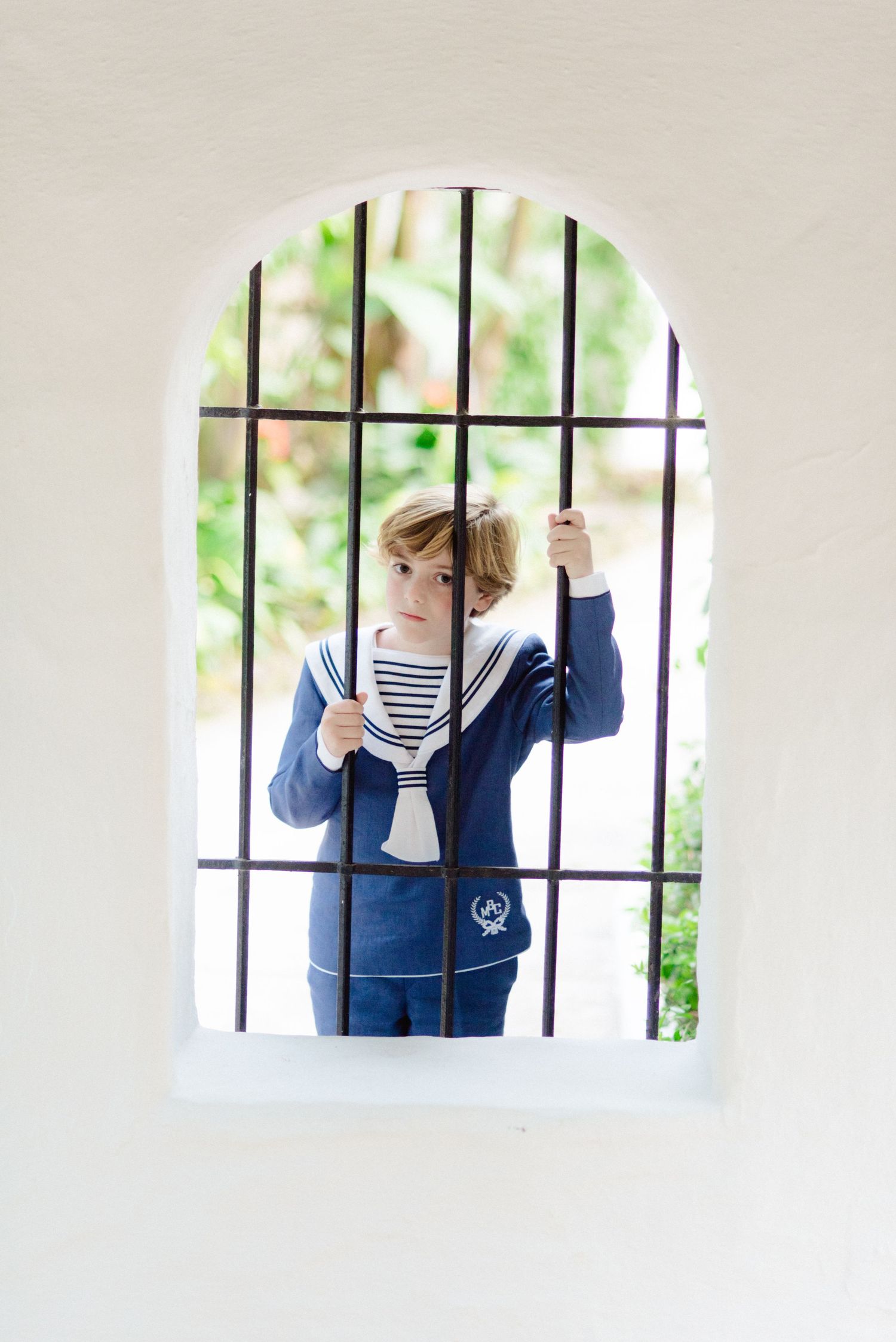 Boy in navy sailor suit standing behind arched iron gate window in white wall.
