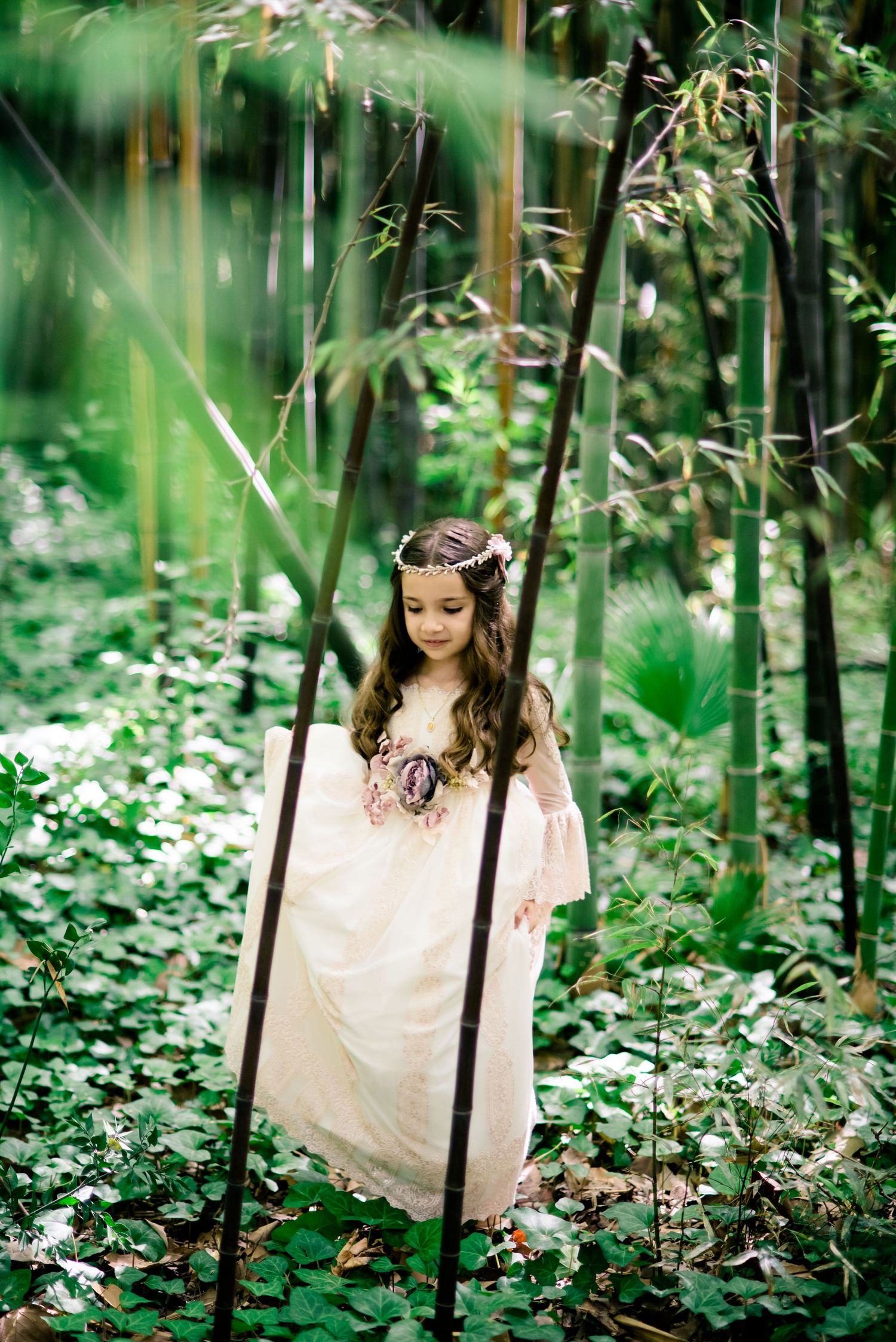 Girl in white lace dress and floral crown walking through lush bamboo and green foliage.