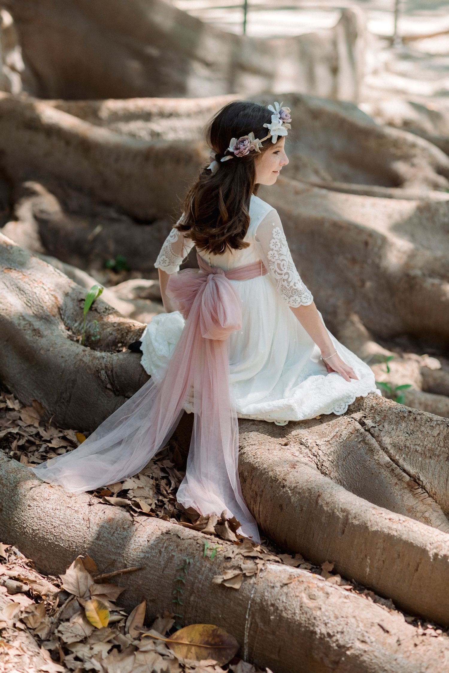Girl in white lace dress with large pink tulle bow sitting on tree roots in garden.