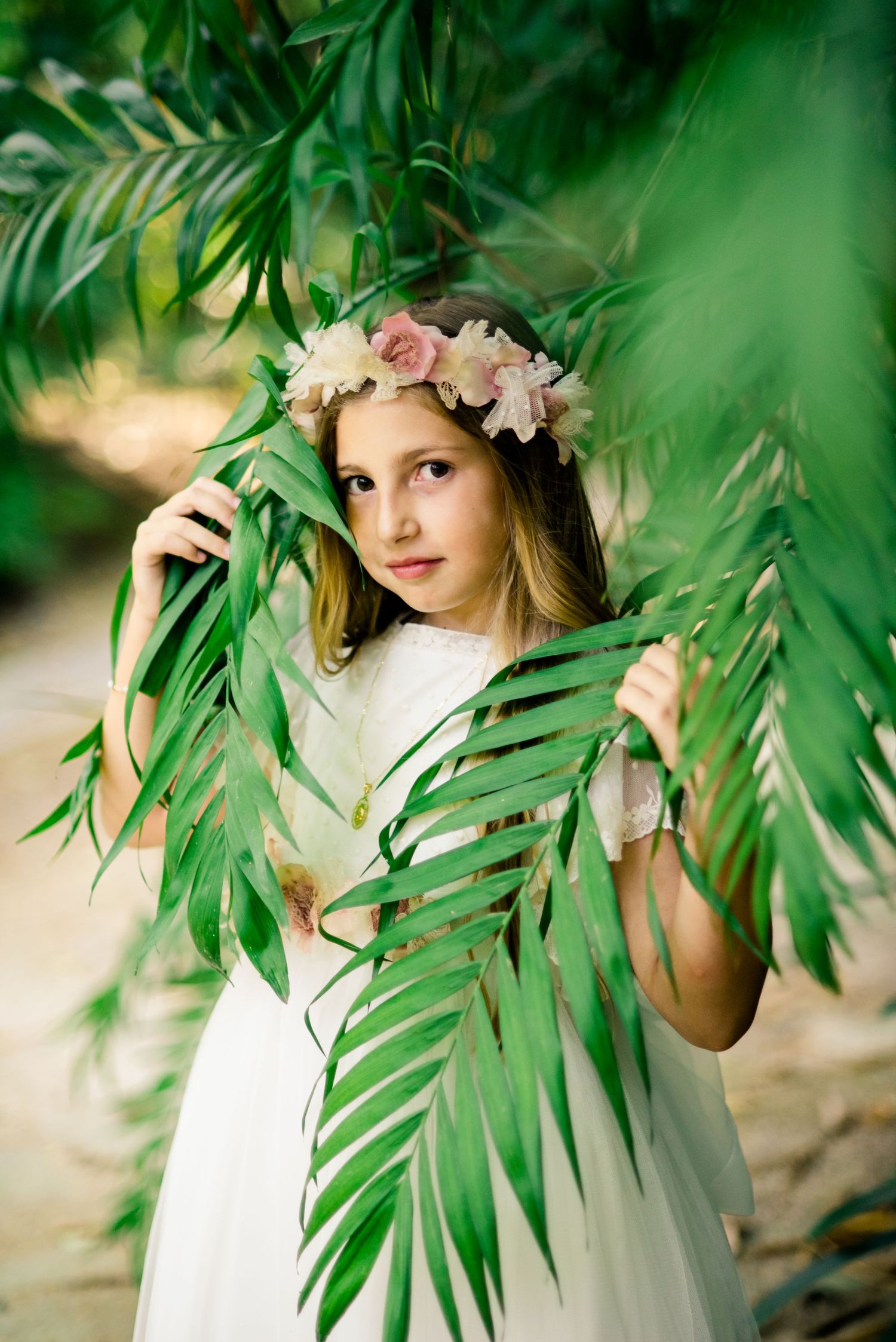 Girl in white communion dress and pink floral crown poses behind tropical palm leaves.