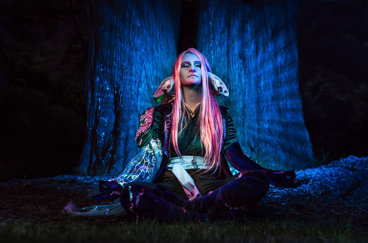 women with long pink hair in elaborate clothing seated meditatively before a glowing tree with blue lighting in a natural setting