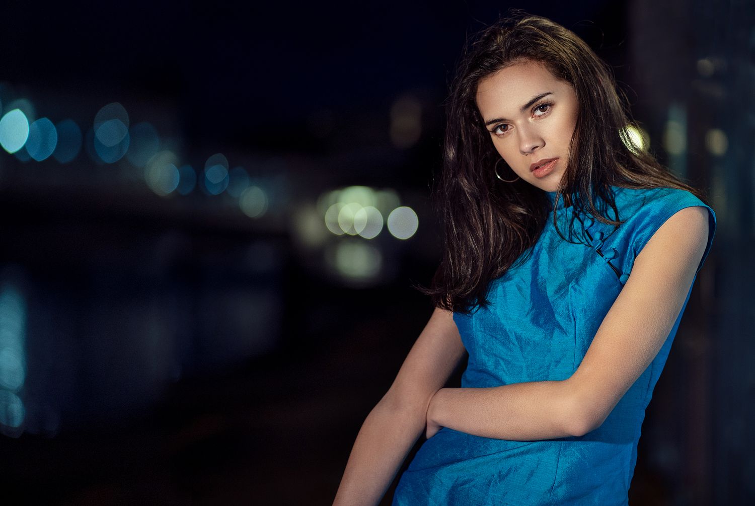 Woman in a blue dress posing confidently at night near a river with soft bokeh lights in the background.