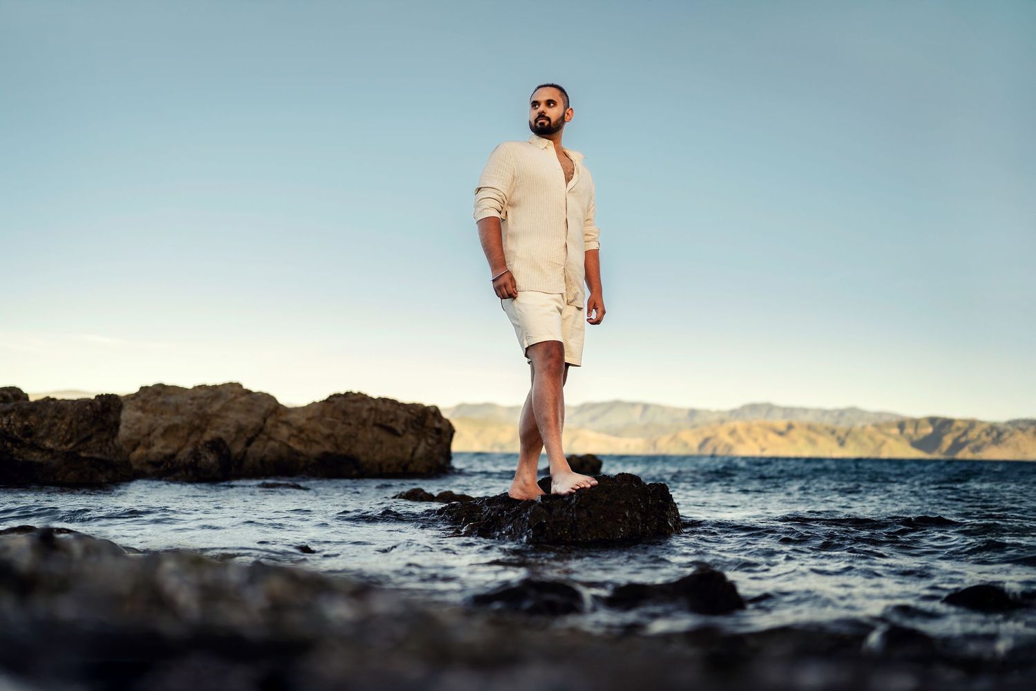 Person standing on rocky coastline during sunset with ocean waves crashing in background.