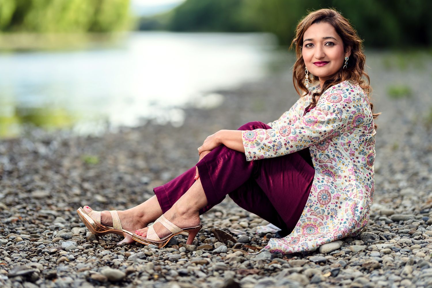 a women sits on pebbles by a river, wearing a colorful patterned shirt and burgundy pants, with sandals. Lush greenery in the background