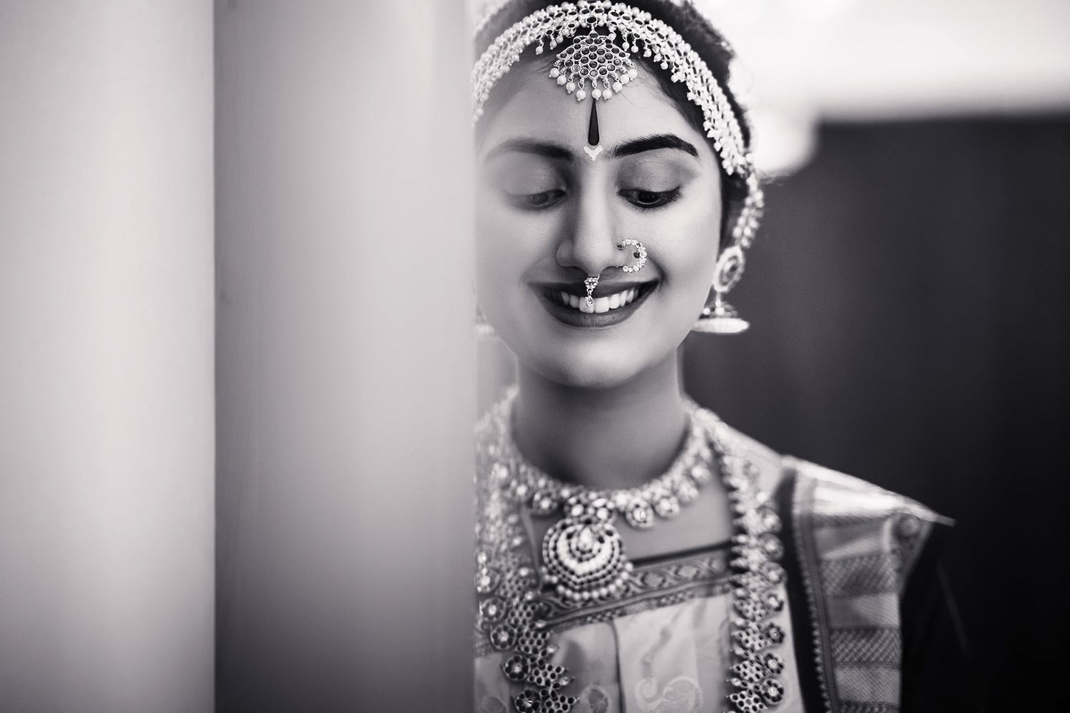 Woman smiling behind a pillar, wearing traditional jewelry and attire, in a black and white photo highlighting her features and accessories.