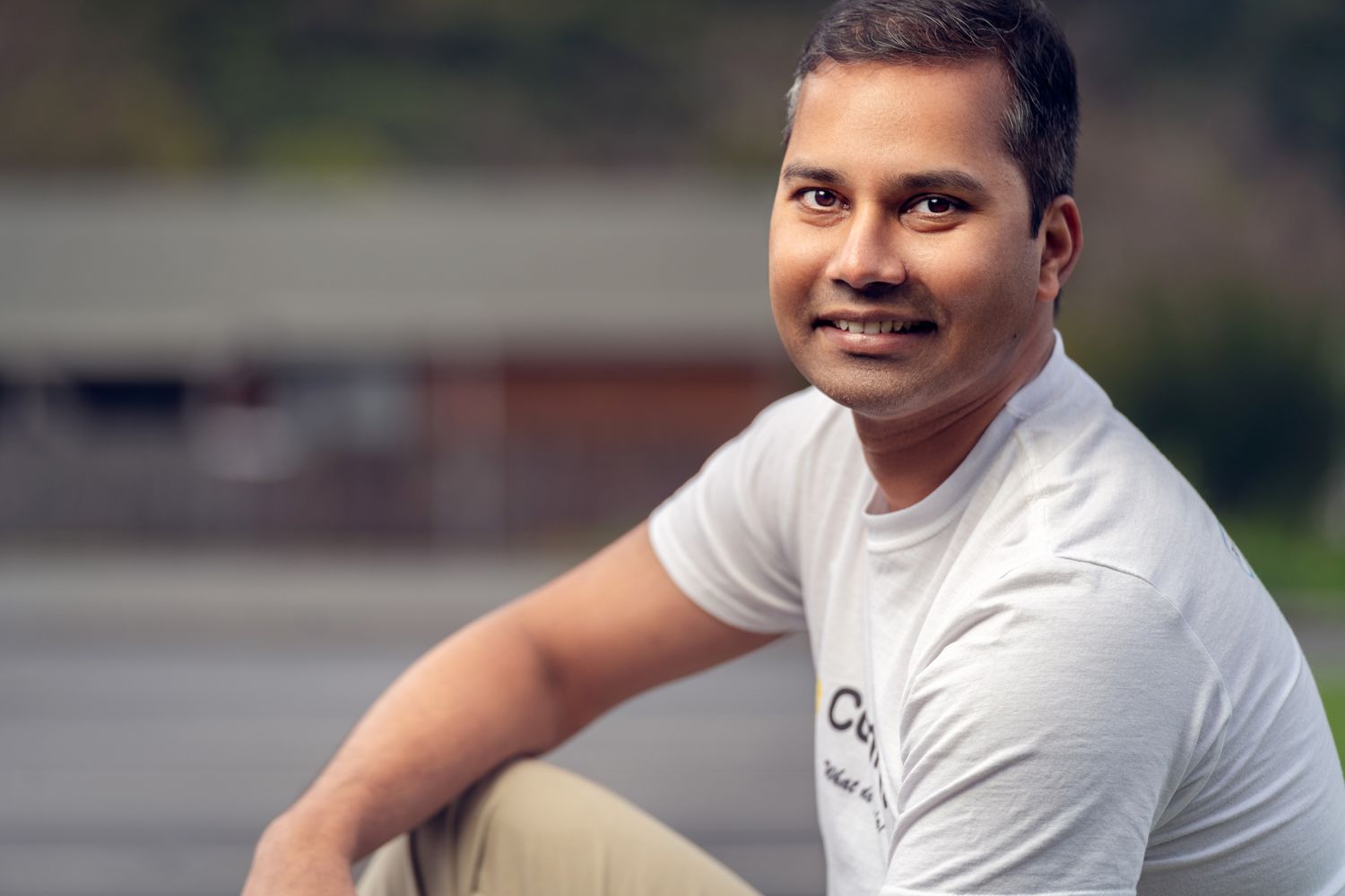Man sitting outdoors in a white t-shirt and light pants, smiling, with blurred greenery and a building in the background.