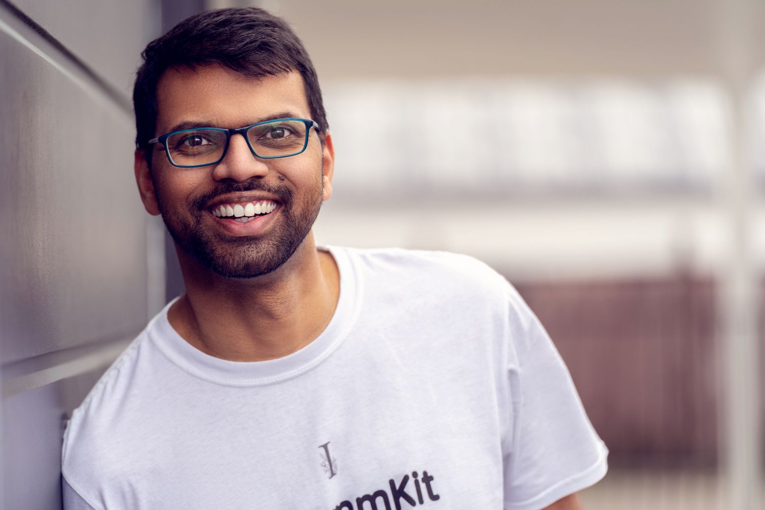 Person wearing glasses and white t-shirt smiles warmly against blurred background.