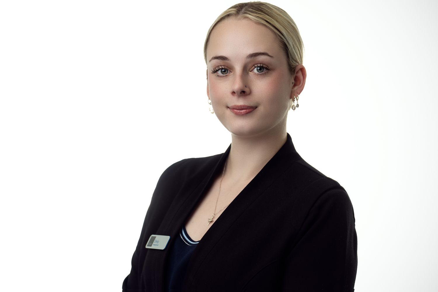 A young woman wearing a black blazer with a name tag, standing confidently with a neutral expression against a white background.