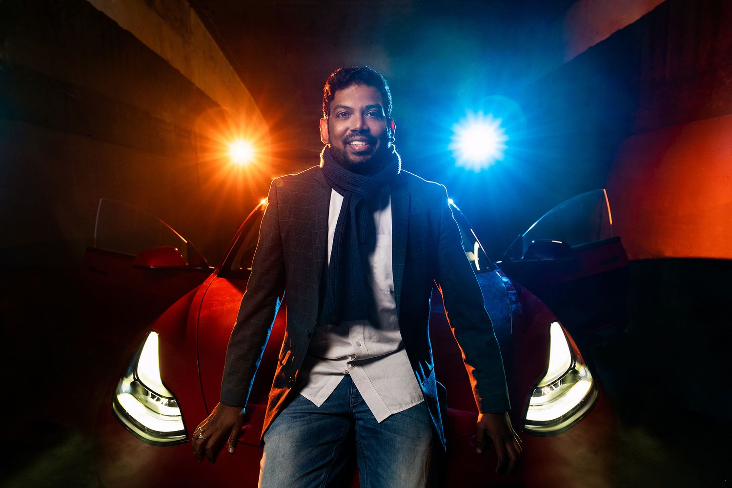 Man in a black blazer and white shirt posing in front of a red car with illuminated headlights and colorful lights in a dimly lit setting.