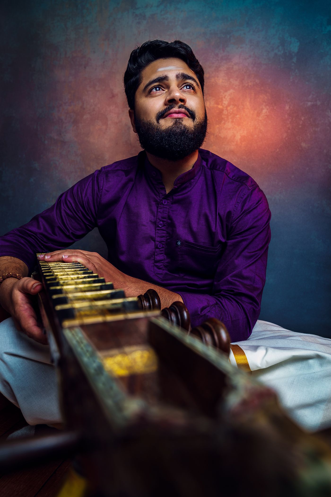 Man in a purple kurta sitting and playing a musical instrument, smiling slightly against a colorful orange and blue background.