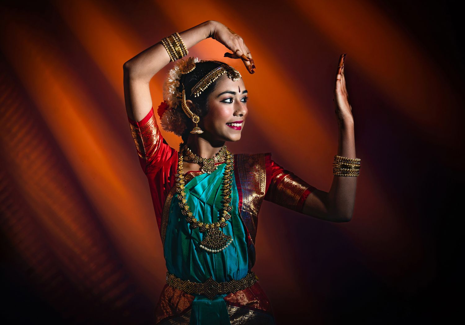 A woman in traditional Indian dance attire strikes a pose, showcasing intricate jewelry and vibrant clothing. The background features warm,