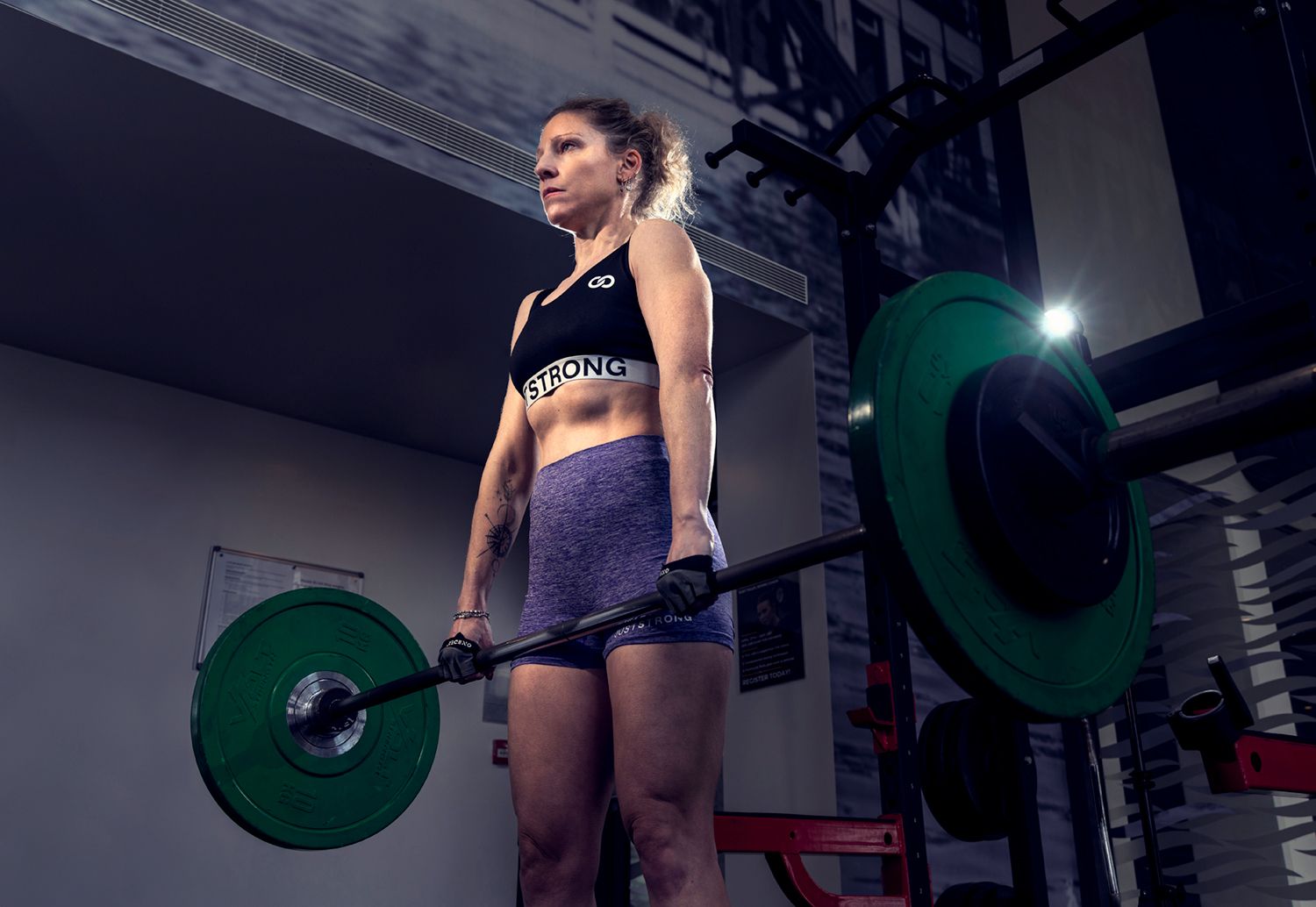 Woman in a black sports bra and purple shorts performing a deadlift in a gym, focused on her form with equipment in the background.