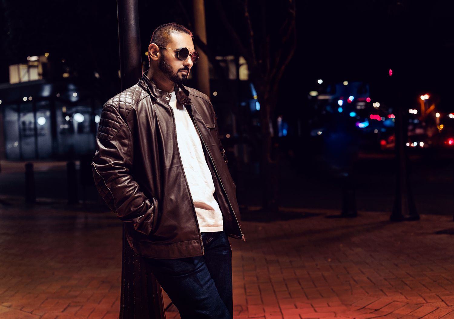 Person leaning against a lamp post at night in a brown leather jacket and white shirt, with city lights in the background.