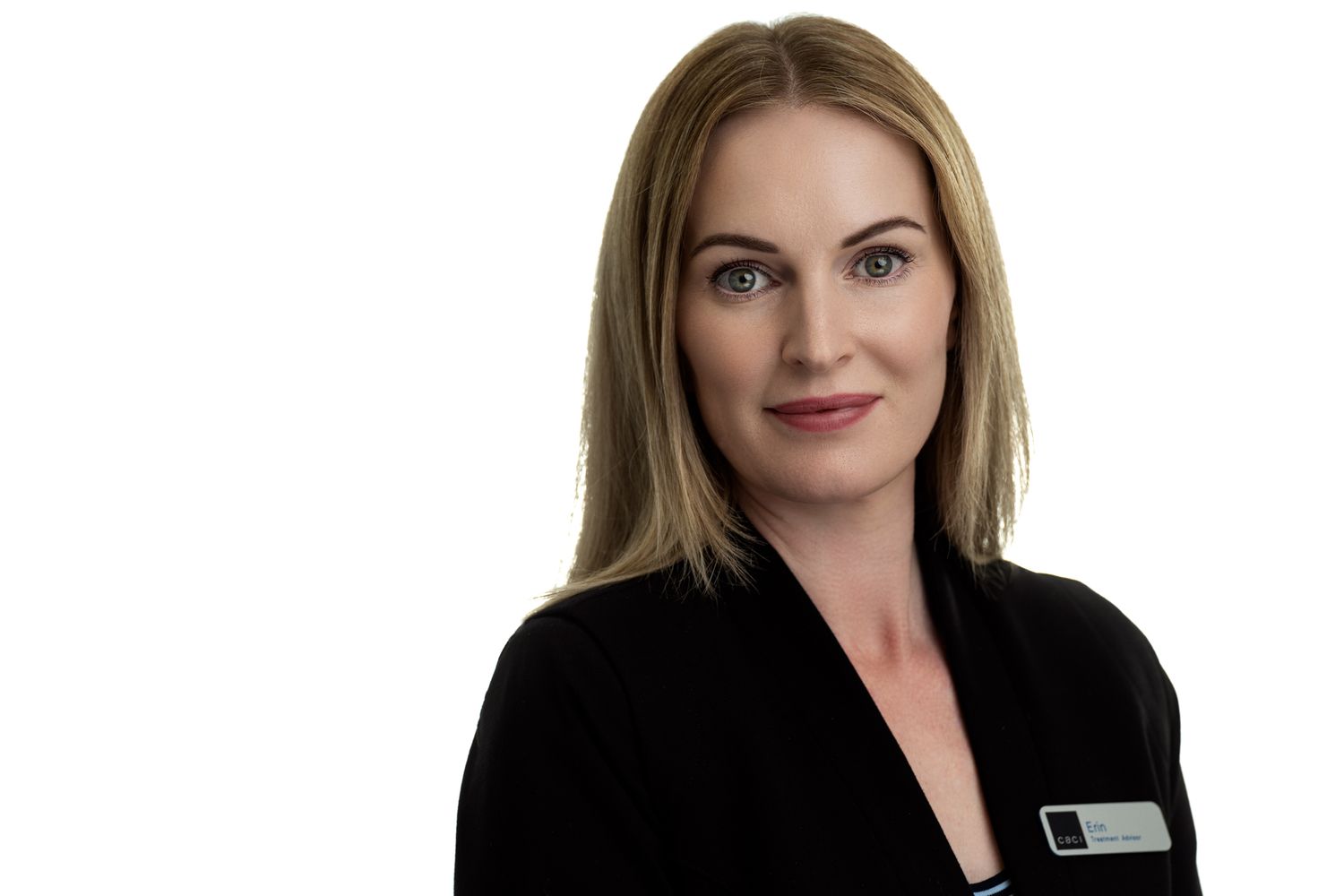 A elder woman wearing a black blazer with a name tag, standing with a neutral expression against a white background.