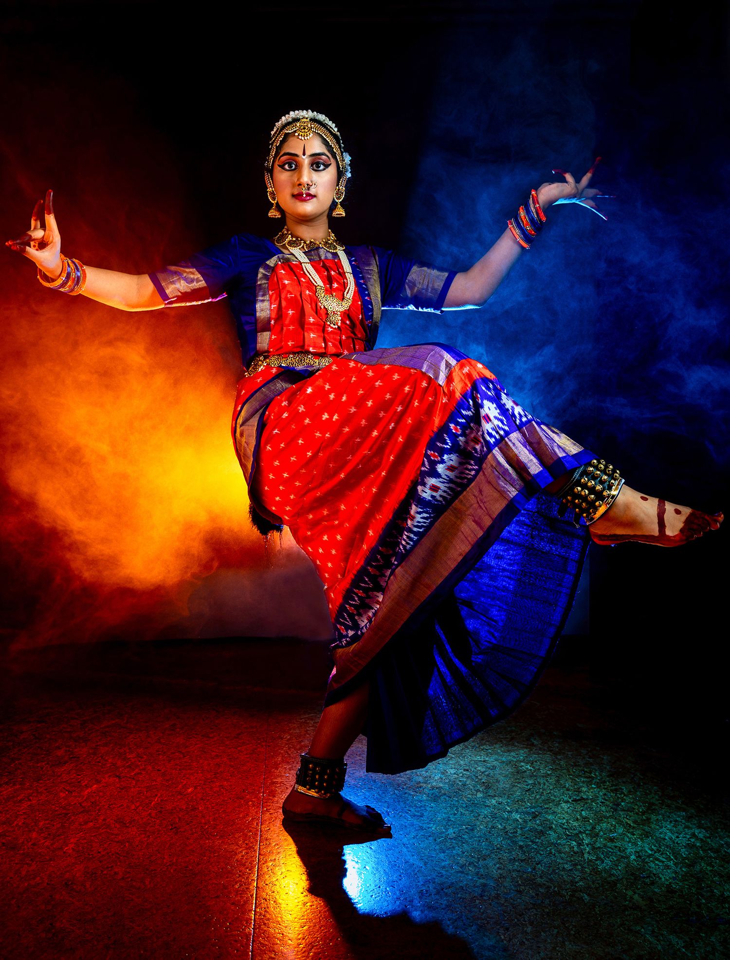 Classical dancer performing on stage in a red and blue costume with jewelry, striking a pose with one leg raised against colorful lights.