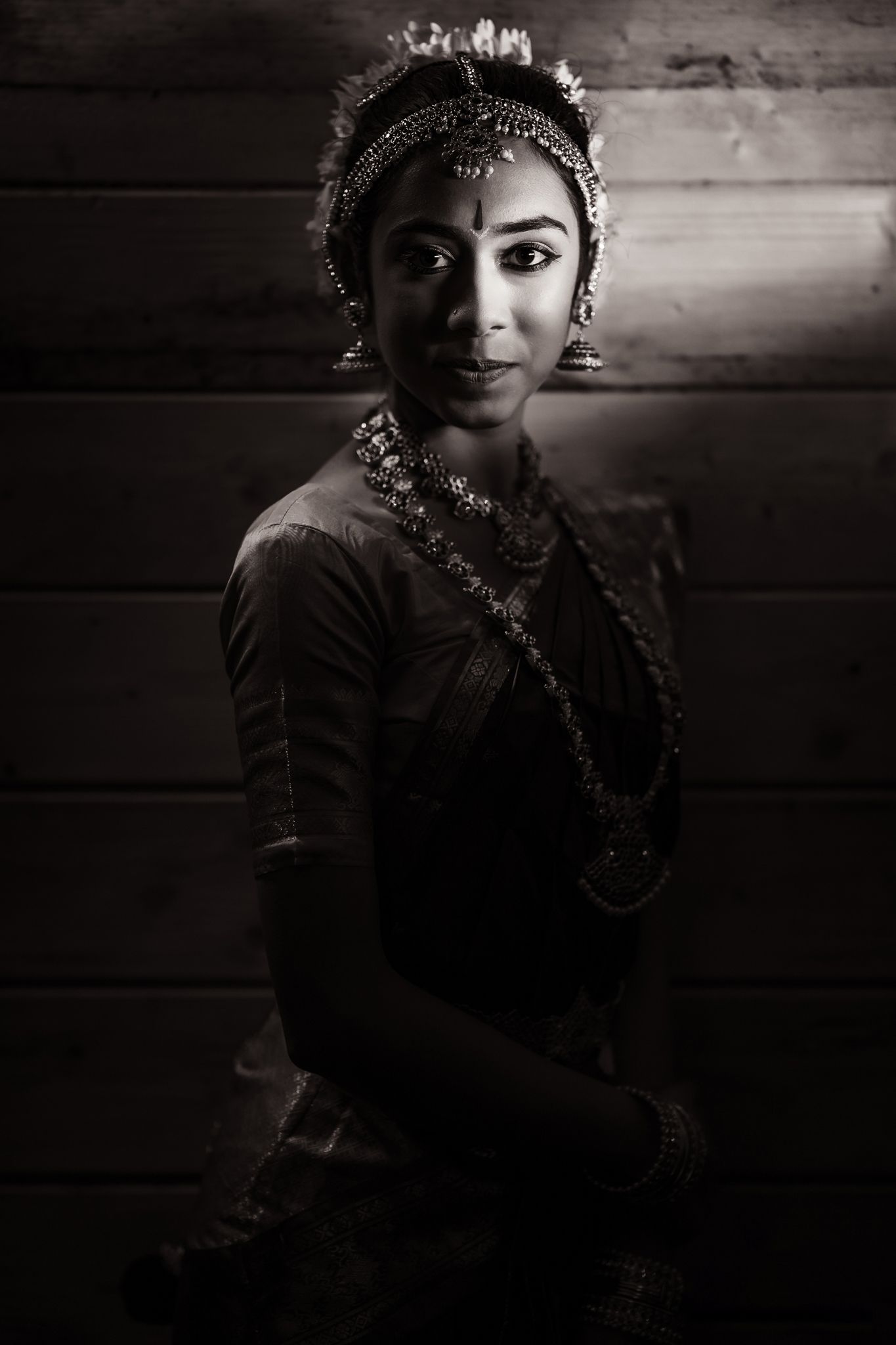 A woman dressed in traditional attire with intricate jewelry, posed against a wooden background. The image is in black and white
