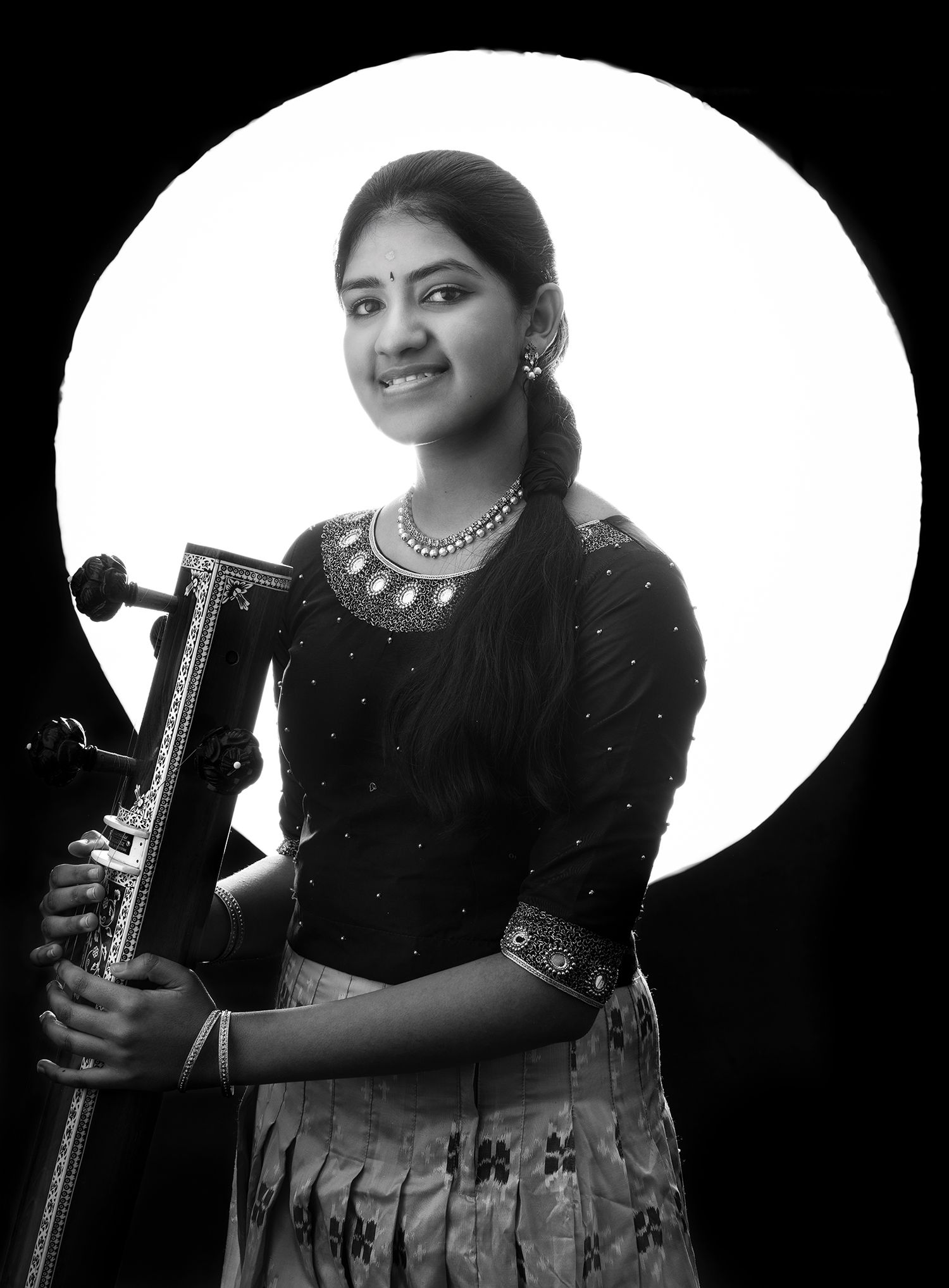 Young girl in traditional attire smiling while holding a musical instrument, with a bright circular light in the black-and-white background.