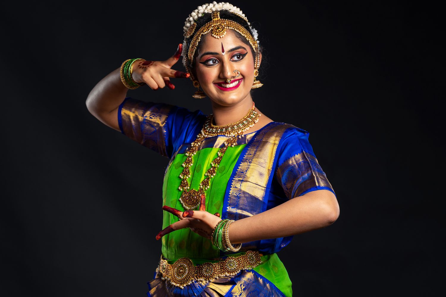 Young woman performing a traditional Indian dance in blue and green attire with jewelry, smiling against a dark background.